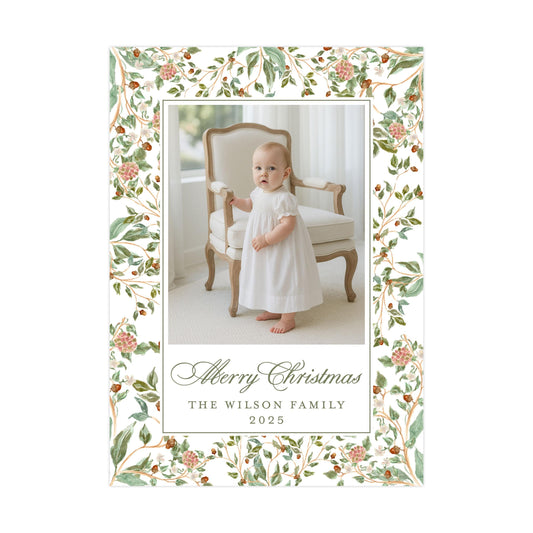 A baby girl in a white dress is standing in a chair with a floral border around the edges of the frame. The text within the frame reads "Merry Christmas" and "The Wilson Family" along with the year "2025".