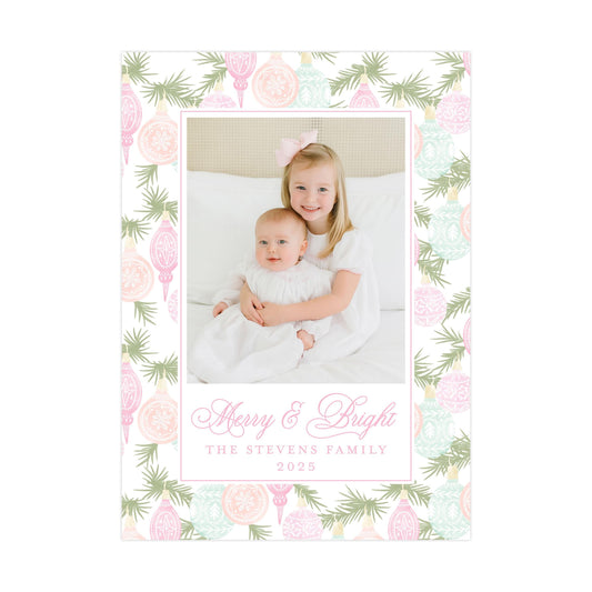 A young girl and a baby are sitting together on a bed, surrounded by a festive floral border. The text "Merry & Bright" and "The Stevens Family" is displayed in a decorative font, and the year "2025" is shown below the image.