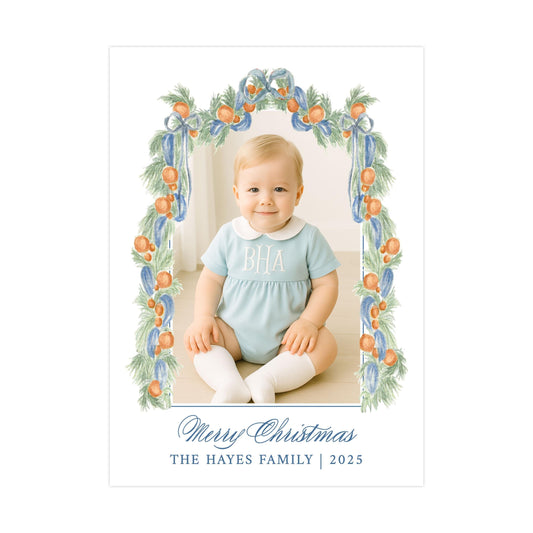 A smiling baby boy wearing a light blue onesie with the initials "BA" on it, sitting on the floor in front of a floral frame. The frame is decorated with orange and green leaves, and the text "Merry Christmas" and "THE HAYES FAMILY | 2025" is displayed below the frame.