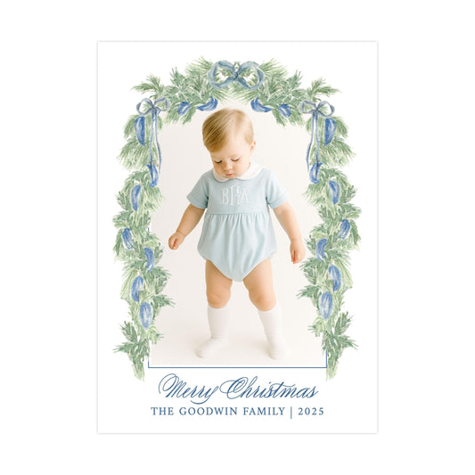 A young child wearing a light blue outfit with a monogrammed "BBA" on the chest, standing in front of a Christmas wreath with blue and green foliage.