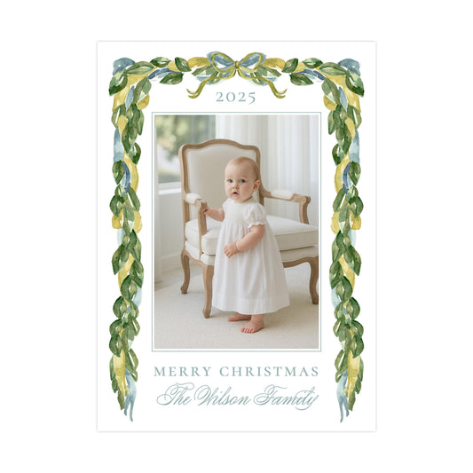 A baby girl in a white dress is standing in a chair, with a decorative border of green leaves and a bow around the top of the frame. The text "Merry Christmas The Wilson Family" is displayed in the image.