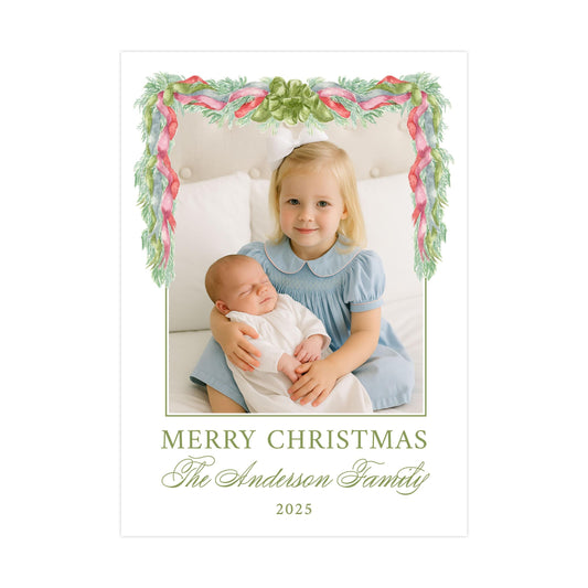 A young girl in a blue dress is holding a sleeping baby in her arms, with a festive Christmas-themed frame surrounding them. The text below the image reads "MERRY CHRISTMAS The Anderson Family 2025".