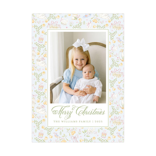 A young girl in a blue dress is holding a baby in a white onesie, both smiling and posing for a Christmas photo. The image is framed with a floral border, and the text "Merry Christmas" and "The Williams Family | 2020" is displayed below the photo.