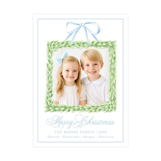 A Christmas card featuring two children, a girl and a boy, smiling in a green wreath frame. The card has a blue ribbon and the text "Merry Christmas" and "The Moore Family | 2025" written on it.