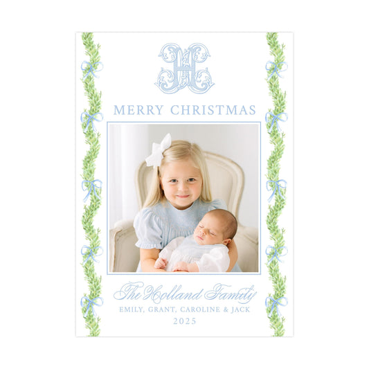 The image is a Christmas card featuring a young girl holding a sleeping baby. The card has a decorative border with green leaves and a monogrammed "M" in the top left corner. The card includes a message that reads "Merry Christmas" and "The Holland Family" along with the names "Emilly, Grant, Caroline & Jack" and the year "2025".