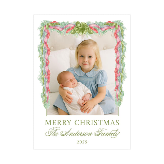 A young girl in a blue dress is holding a sleeping baby in her arms, with a festive Christmas-themed frame surrounding them. The text below the image reads "MERRY CHRISTMAS The Anderson Family 2025".
