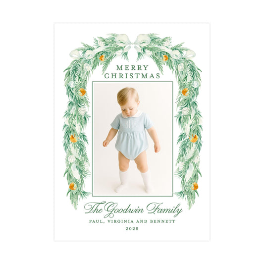 A Christmas card featuring a baby in a light blue outfit, surrounded by a green wreath with orange flowers. The card has a message that reads "Merry Christmas The Goodwin Family Paul, Virginia and Bennett 2005".