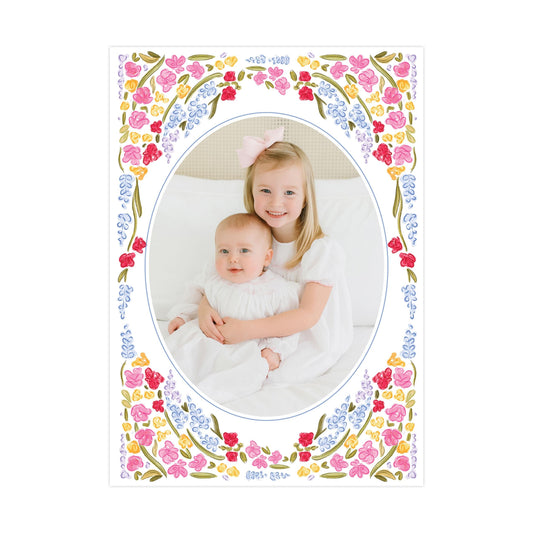 A young girl in a white dress is holding a baby in a white dress, both smiling and looking at the camera. The image is framed by a decorative border with colorful flowers.