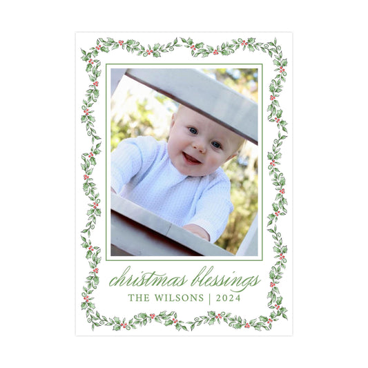 A smiling baby in a white outfit peeking out from behind a decorative frame with a holly leaf border.