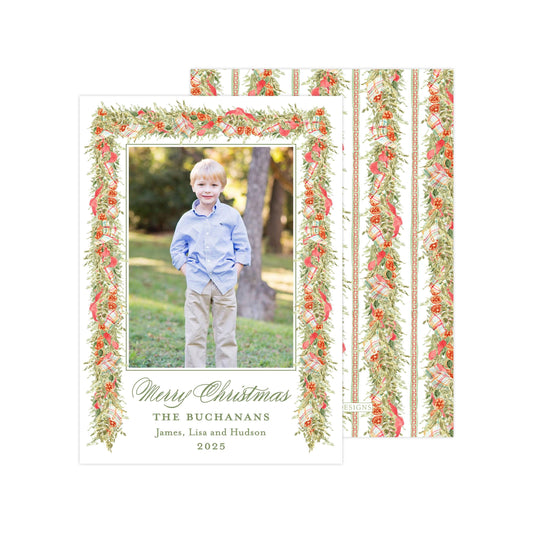 A young boy is standing in a grassy area, smiling and posing for a Christmas card. The card has a floral border and a photo of the boy in the center. The text on the card reads "Merry Christmas THE BUCKANNS James, Liza and Hudson 2020".