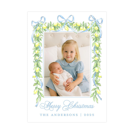 A young girl in a blue dress is holding a sleeping baby in her arms, with a decorative frame around them. The frame has a blue border and green leaves, and the text &quot;Merry Christmas&quot; and &quot;THE ANDERSons | 2025&quot; is displayed below the frame.