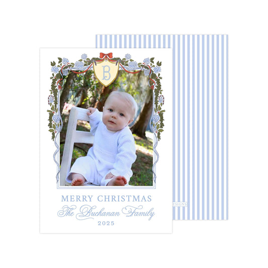 A baby sitting in a chair, surrounded by a decorative border with a blue and white striped pattern. The baby is wearing a white outfit. The image is framed with a border that includes a floral design and a shield-shaped emblem. The text at the bottom of the image reads "MERRY CHRISTMAS The Buchanan Family 2015".