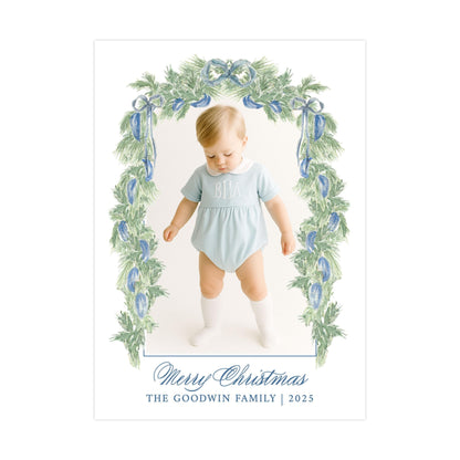 A young child wearing a light blue outfit with a monogrammed "BBA" on the chest, standing in front of a Christmas wreath with blue and green foliage.