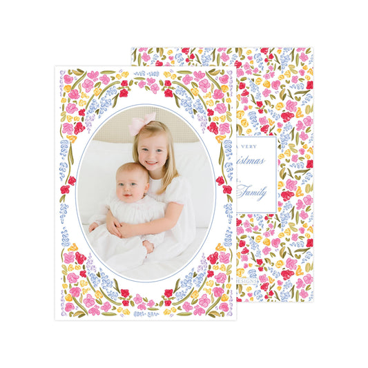 A young girl in a white dress is holding a baby in a white onesie, both smiling and looking at the camera. The background is decorated with a floral pattern in various colors.