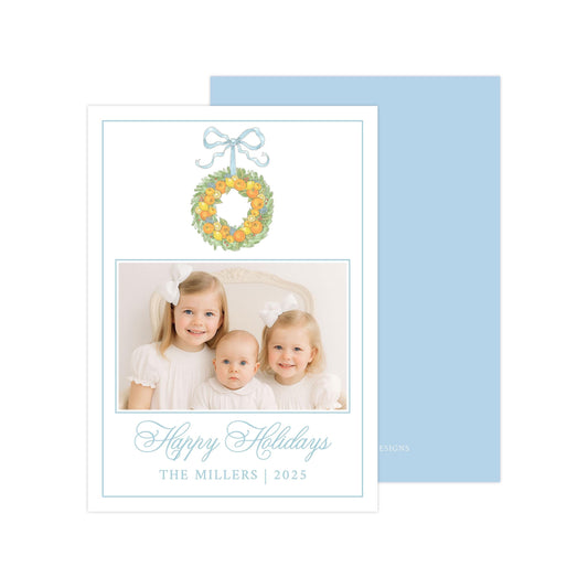 A family photo of three young girls, likely sisters, smiling together in front of a wreath. The photo is framed with a blue border and the text "Happy Holidays The Millers | 2025" is displayed below the image.