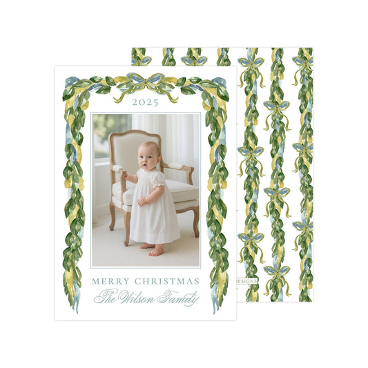 A white card with a photo of a baby girl in a white dress, surrounded by green and yellow ribbons. The card has a "Merry Christmas" message and the name "The Wilson Family" written on it.