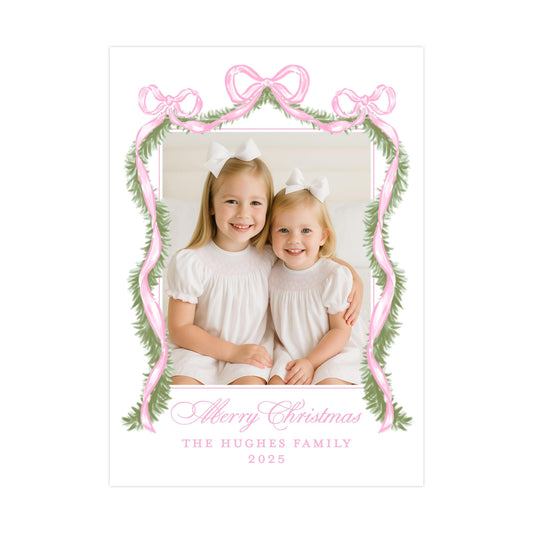 A photo of two young girls smiling and sitting together, framed by a pink ribbon and greenery, with the text &quot;Merry Christmas&quot; and &quot;The Hughes Family&quot; below.