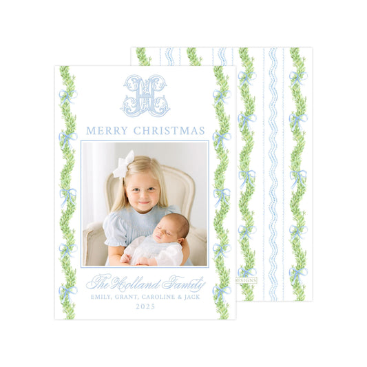 A Christmas card featuring a young girl holding a baby, with a decorative border of green and blue leaves. The card has a message that reads &quot;Merry Christmas&quot; and includes the names &quot;Emilly, Grant, Caroline & Jack&quot; along with the year &quot;2025&quot;.