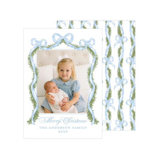 A young girl is holding a sleeping baby in a cozy setting, surrounded by a festive wreath and ribbons. The image is a holiday-themed card with a personalized message and a family photo.