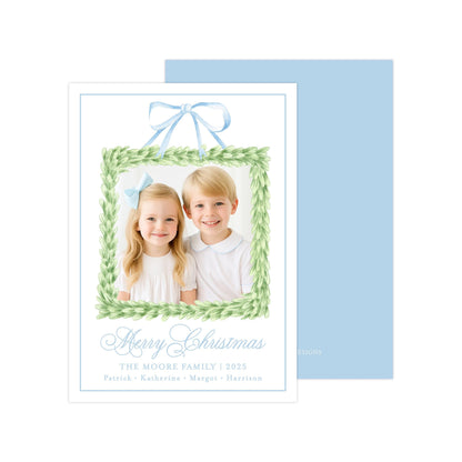 A Christmas card featuring two children, a girl and a boy, smiling in front of a green wreath frame. The card has a blue background and a blue ribbon bow.