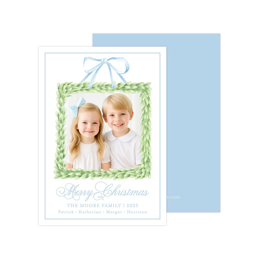 A Christmas card featuring two children, a girl and a boy, smiling in front of a green wreath frame. The card has a blue background and a blue ribbon bow.