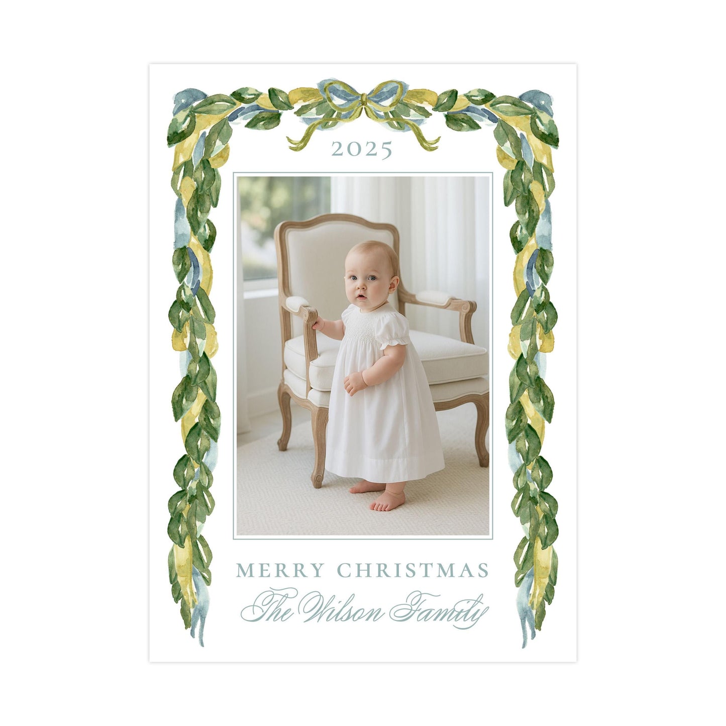 A baby girl in a white dress is standing in a chair, with a decorative border of green leaves and a bow around the top of the frame. The text "Merry Christmas The Wilson Family" is displayed in the image.