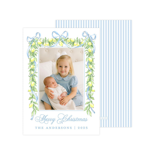 A young girl in a blue dress is holding a sleeping baby in her arms, with a framed photo of them in front of a decorative border of green leaves and blue ribbons. The text &quot;Merry Christmas&quot; and &quot;The Andersons | 2025&quot; is displayed below the photo.