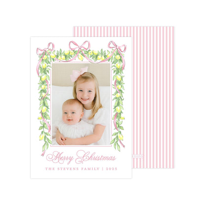 A Christmas card featuring two young girls, one wearing a white dress and the other in a pink dress, smiling and embracing each other. The card has a pink and white striped border and a floral design with lemons and ribbons. The text on the card reads &quot;Merry Christmas&quot; and &quot;The Stevens Family | 2020&quot;.