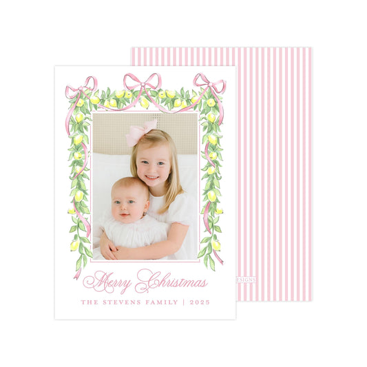 A Christmas card featuring two young girls, one wearing a white dress and the other in a pink dress, smiling and embracing each other. The card has a pink and white striped border and a floral design with lemons and ribbons. The text on the card reads &quot;Merry Christmas&quot; and &quot;The Stevens Family | 2020&quot;.