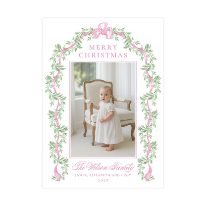 A baby dressed in a white gown is standing in front of a chair, with a floral border and the text &quot;Merry Christmas&quot; above the photo.