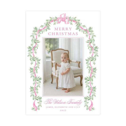 A baby dressed in a white gown is standing in front of a chair, with a floral border and the text &quot;Merry Christmas&quot; above the photo.