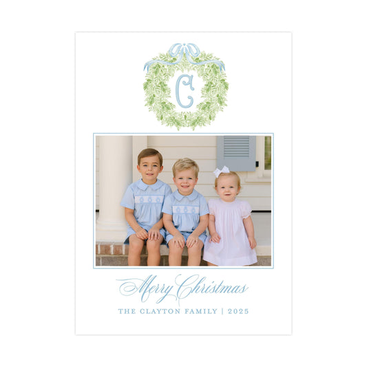 A family photo of three children, two boys and one girl, sitting on a porch with a wreath above them. The text below the photo reads &quot;Merry Christmas&quot; and &quot;THE CLAYTON FAMILY | 2025&quot;.