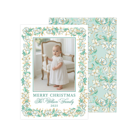 A baby dressed in a white gown is standing in front of a chair, with a floral patterned background behind her. The text on the card reads "Merry Christmas The Wilson Family 2025".