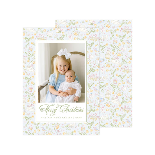 A young girl in a blue dress is holding a baby in a white onesie, both smiling and posing for a Christmas card. The card has a floral pattern and the text "Merry Christmas" and "The Williams Family" along with the year "2015".