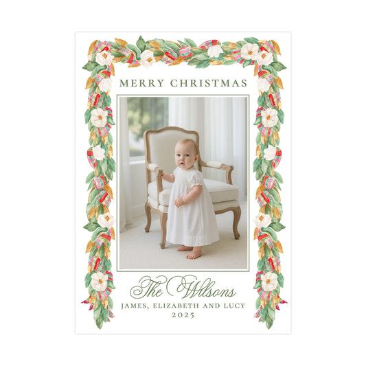 A framed photograph of a baby girl in a white dress, standing in front of a chair with a floral border. The text &quot;MERRY CHRISTMAS&quot; is displayed above the photo, and &quot;The Wiltsons&quot; is written below the photo.