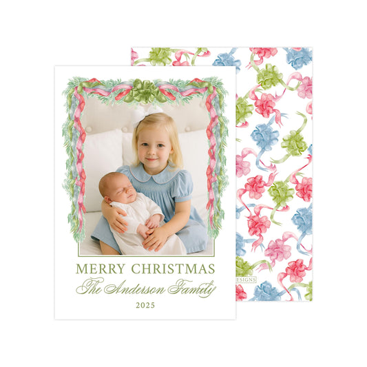 A young girl in a blue dress is holding a baby in her arms, with a floral wreath frame surrounding them. The text "MERRY CHRISTMAS The Anderson Family 2020" is displayed below the image.