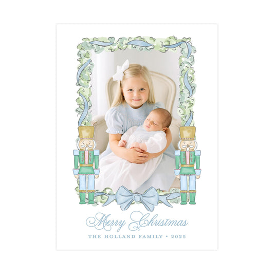A young girl is holding a baby in her arms, with a nutcracker figure on either side of the frame. The image is framed with a decorative border and the text &quot;Merry Christmas&quot; and &quot;The Holland Family&quot; is displayed below.