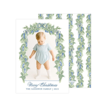 A photo of a baby wearing a light blue onesie, standing in front of a floral archway with a "Merry Christmas" greeting.