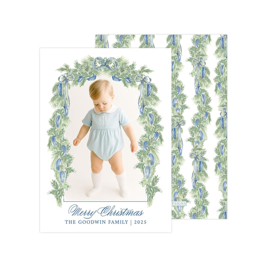 A photo of a baby wearing a light blue onesie, standing in front of a floral archway with a "Merry Christmas" greeting.