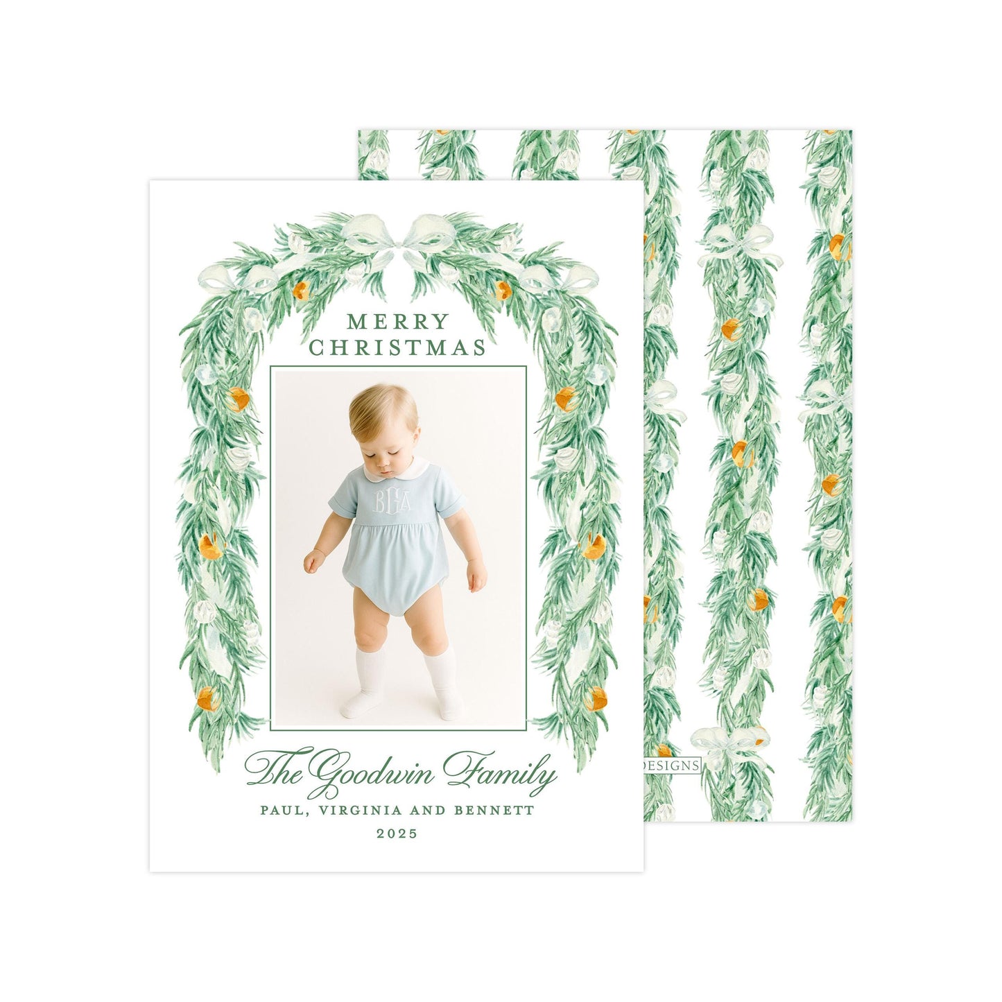 A Christmas card featuring a baby wearing a light blue outfit, surrounded by a green wreath with white flowers and orange ornaments. The card has a festive design with a green border and text that reads "Merry Christmas" and "The Goodwin Family".