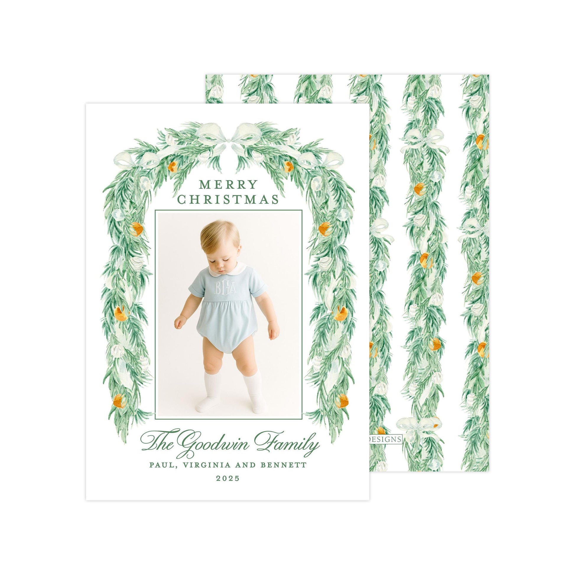 A Christmas card featuring a baby wearing a light blue outfit, surrounded by a green wreath with white flowers and orange ornaments. The card has a festive design with a green border and text that reads "Merry Christmas" and "The Goodwin Family".