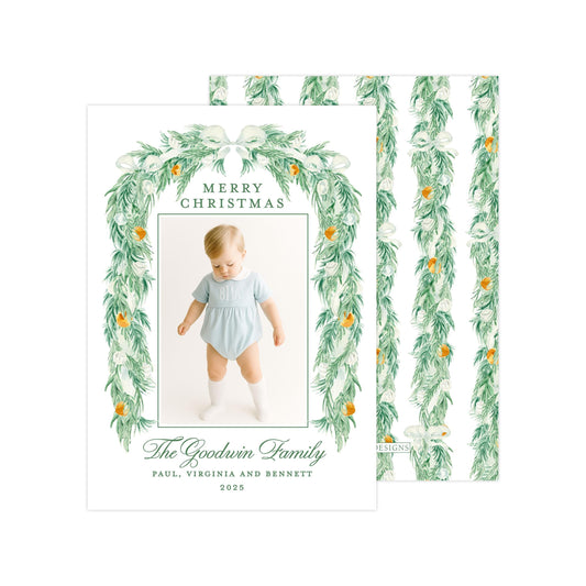 A Christmas card featuring a baby wearing a light blue outfit, surrounded by a green wreath with white flowers and orange ornaments. The card has a festive design with a green border and text that reads "Merry Christmas" and "The Goodwin Family".