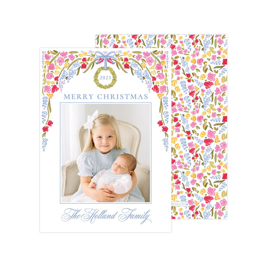 A Christmas card featuring a young girl holding a sleeping baby, with a floral wreath design above the photo.