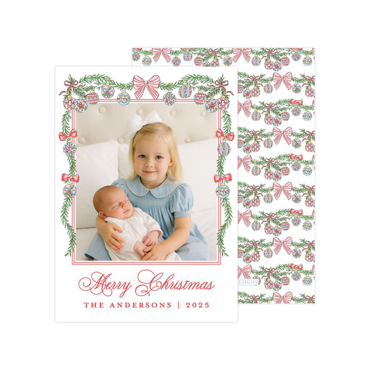 A young girl in a blue dress is holding a sleeping baby in her arms, with a festive Christmas-themed background featuring red and green decorations.