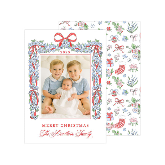 A Christmas card featuring two young boys sitting on a bed, with a baby between them, and a floral border around the photo. The card has a festive design with red and white ribbons and flowers, and the text &quot;Merry Christmas The Bradbury Family&quot; is displayed.