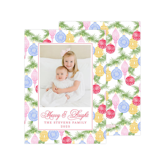 A photo of two young girls, one wearing a white dress and the other in a white dress, sitting together on a bed with Christmas ornaments on the bedspread. The image is framed with a border of Christmas ornaments and greenery, and the text &quot;Merry & Bright&quot; and &quot;THE STEVENS FAMILY&quot; is displayed below the photo.