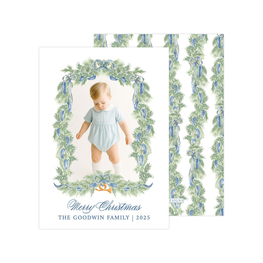 A photo of a baby wearing a blue onesie, surrounded by a decorative frame with a wreath of greenery and blue ribbons. The text on the card reads "Merry Christmas THE GOODWIN FAMILY | 2015".